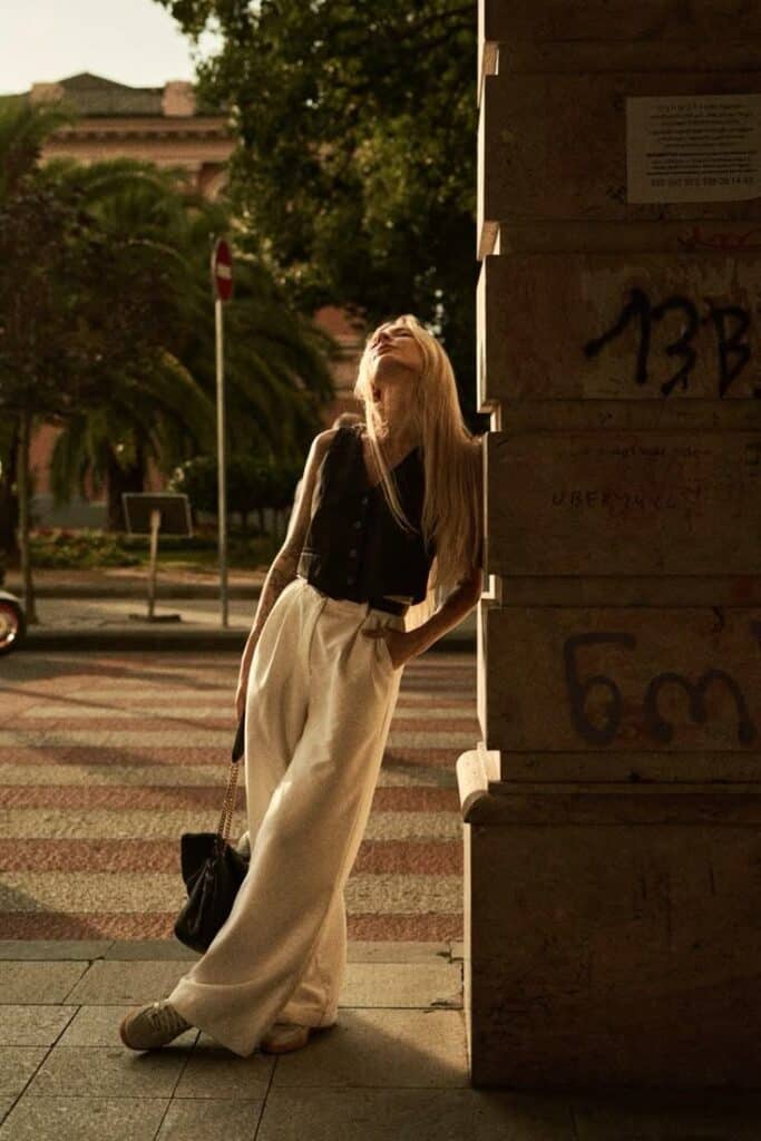 photo Woman in stylish black vest and white pants leans against a textured wall in sunny urban setting, casting a relaxed pose. | Sky Rye Design Woman in stylish black vest and white pants leans against a textured wall in sunny urban setting, casting a relaxed pose.