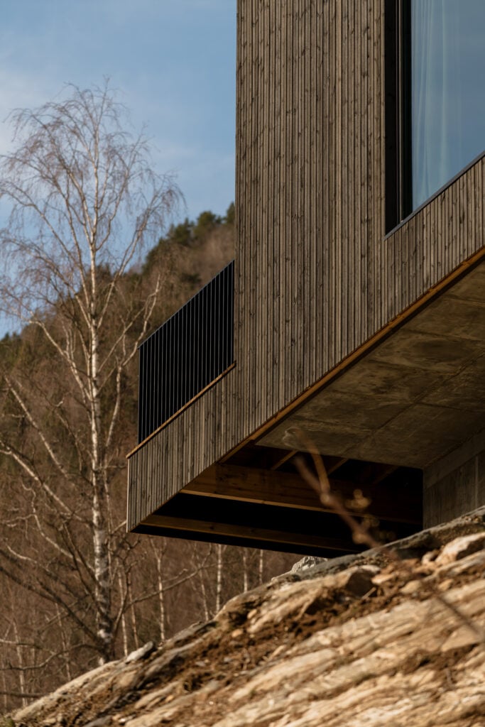 Modern wood-clad house design with large windows, set against a forested hillside background. Serene and minimalist architecture.