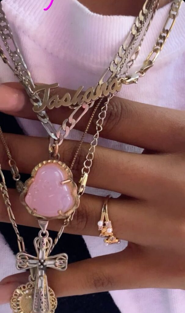 shanadenai Close-up of layered gold necklaces and rings, featuring a nameplate, pink pendant, cross, and pearl-adorned jewelry on a person’s hand. | Sky Rye Design Close-up of layered gold necklaces and rings, featuring a nameplate, pink pendant, cross, and pearl-adorned jewelry on a person’s hand.