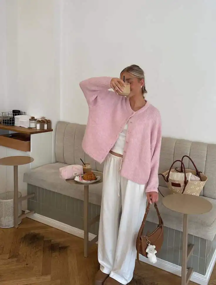 Woman in cozy pink cardigan and white pants enjoying a coffee in a chic cafe setting, with pastries and handbag on side table.