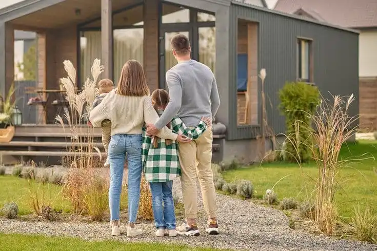 Family standing outside a modern house, embracing and looking at the home, surrounded by a lush garden landscape.