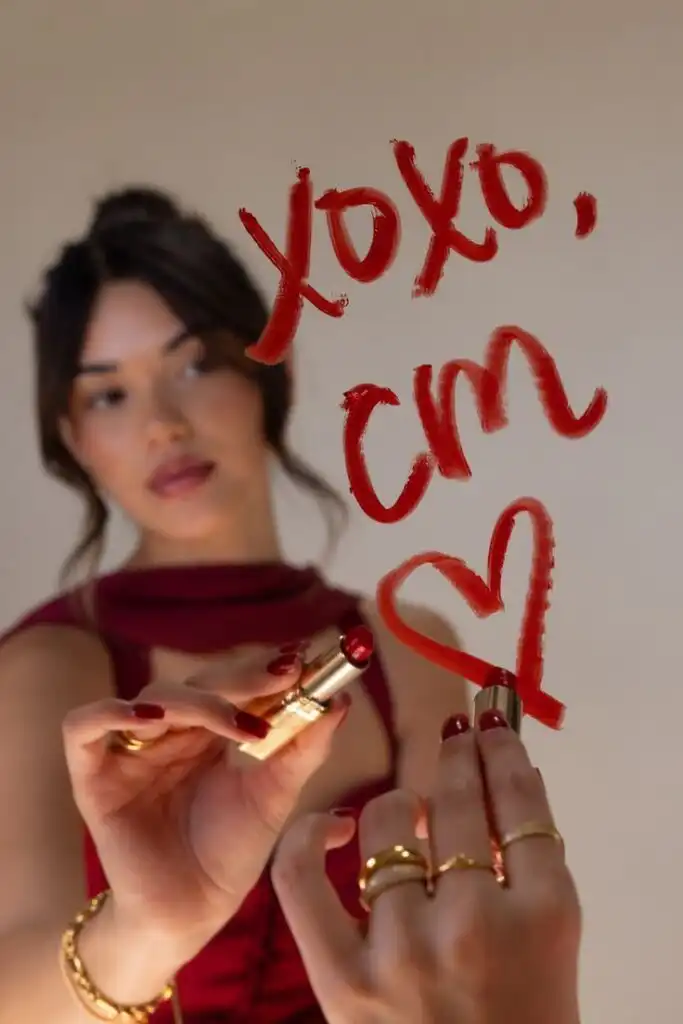 _ Woman writing xoxo and heart on mirror with lipstick, wearing red outfit and gold rings, enhancing beauty and style. | Sky Rye Design Woman writing xoxo and heart on mirror with lipstick, wearing red outfit and gold rings, enhancing beauty and style.