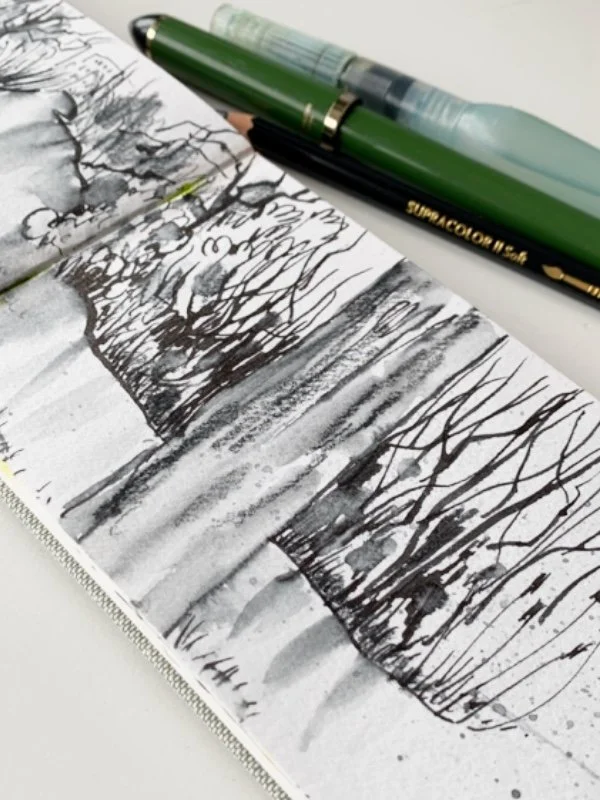 Artistic sketch of trees in a notebook with a green brush pen and pencil placed on top, capturing nature with abstract lines and shades.