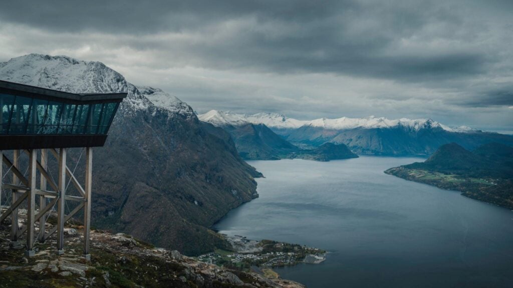 Scenic view of a snowy mountain range and fjord, with a modern viewing platform jutting out over a rocky landscape under a cloudy sky.