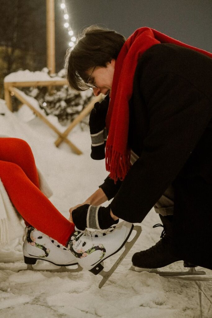 Парная фотосессия на катке _ Зимняя эстетика Person kneeling on snowy ground helps tie ice skates, wearing red scarf and gloves, enhancing winter scene ambiance with fall lights. | Sky Rye Design Person kneeling on snowy ground helps tie ice skates, wearing red scarf and gloves, enhancing winter scene ambiance with fall lights.