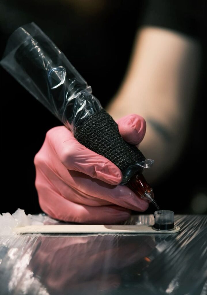Серьезные люди в розовых перчатках Tattoo artist in pink gloves prepares tattoo machine with ink cup on table, highlighting precision in art. | Sky Rye Design Tattoo artist in pink gloves prepares tattoo machine with ink cup on table, highlighting precision in art.