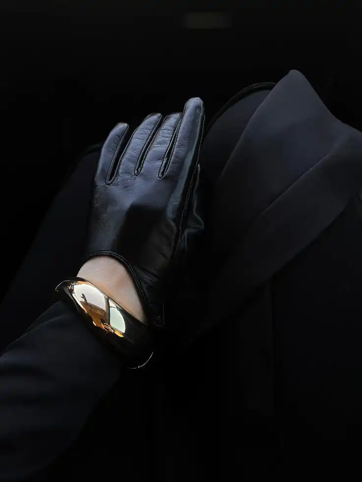 Elegant black leather glove rests on lap with a shiny gold bracelet, reflecting light in a dark, sophisticated setting.