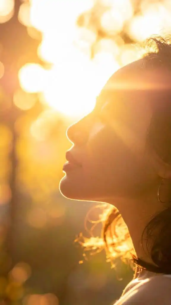 Woman enjoying golden hour sunshine with eyes closed, peaceful and serene, sun rays illuminating her face, nature background.