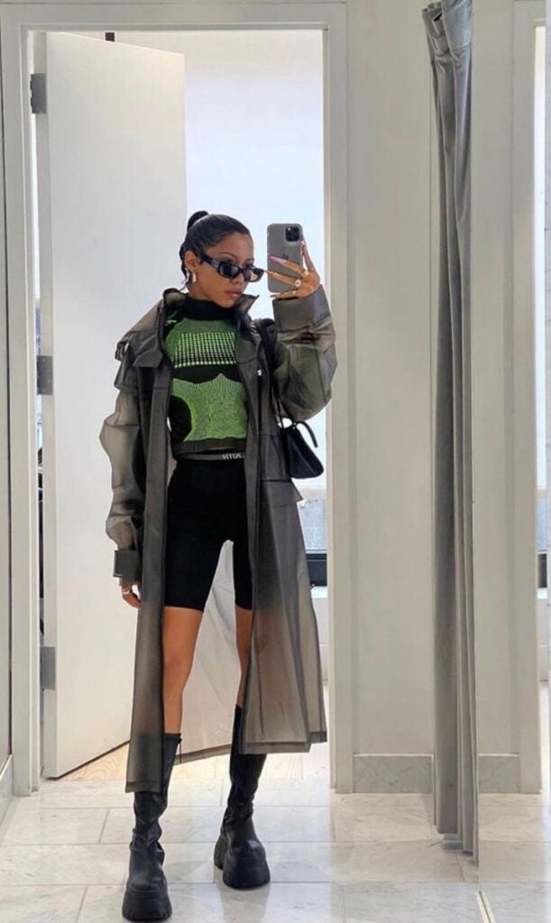 🌮 Fashionable woman in long coat, green top, black shorts, and boots takes a stylish mirror selfie in a modern room. | Sky Rye Design Fashionable woman in long coat, green top, black shorts, and boots takes a stylish mirror selfie in a modern room.