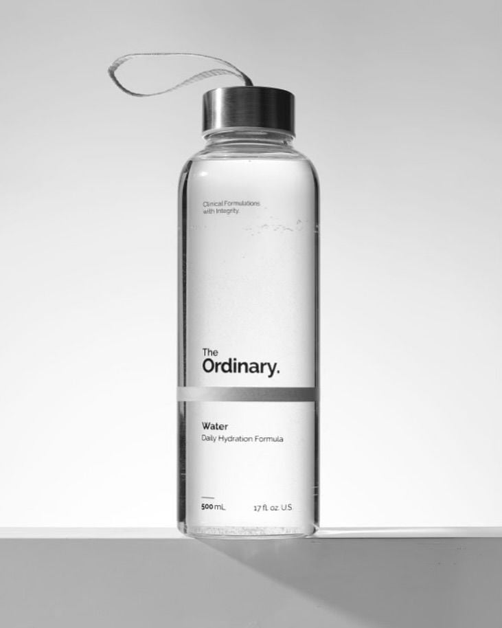 Product shot of The Ordinary Water (500 mL) clear bottle — minimalist hydration formula with metal cap and wrist strap on pale background.