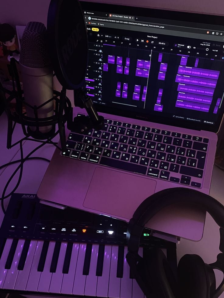 Home music studio setup: laptop with DAW showing purple waveforms, condenser microphone, MIDI keyboard and over-ear headphones.