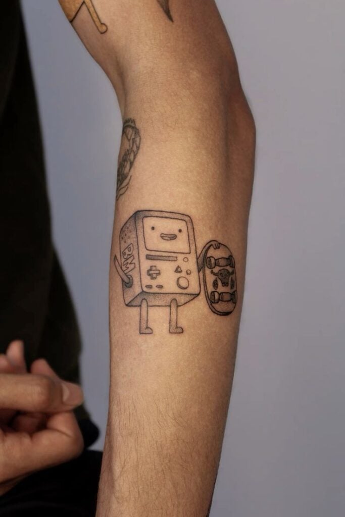 Forearm tattoo of BMO-style handheld game console character holding a skateboard, simple black linework cartoon tattoo on inner forearm.