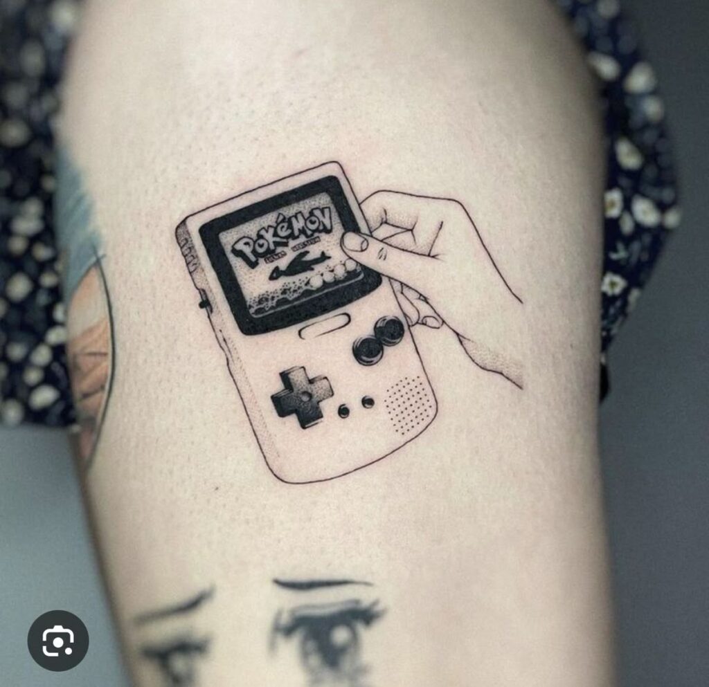 Black linework tattoo of a hand holding a classic Game Boy displaying Pokémon on screen, minimal dot shading on upper thigh.
