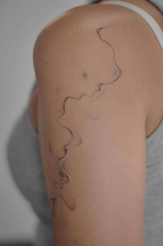 Close-up of minimalist fine-line arm tattoo: abstract black swirl with tiny star accents on woman's upper arm in sleeveless grey top