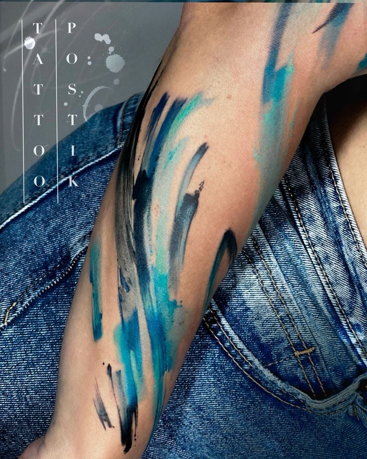 Forearm watercolor tattoo: abstract blue and black brushstrokes over denim, modern minimalist arm art.