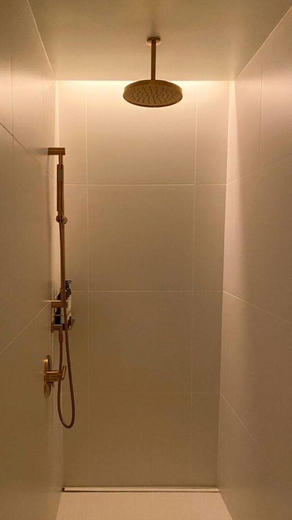 Minimalist modern tiled shower with ceiling-mounted rainfall showerhead and matte gold hand shower fixtures, warm recessed lighting.