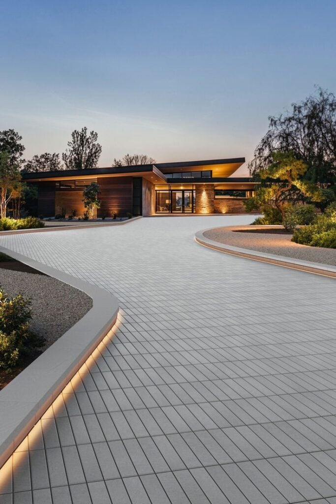 Contemporary luxury house with curved paved driveway, minimalist landscaping and warm exterior lighting at dusk