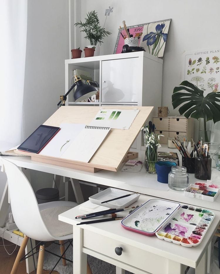 Bright home artist workspace with angled drawing board, watercolor palette and brushes, tablet, desk lamp, plants and botanical prints. Guide to Relocating