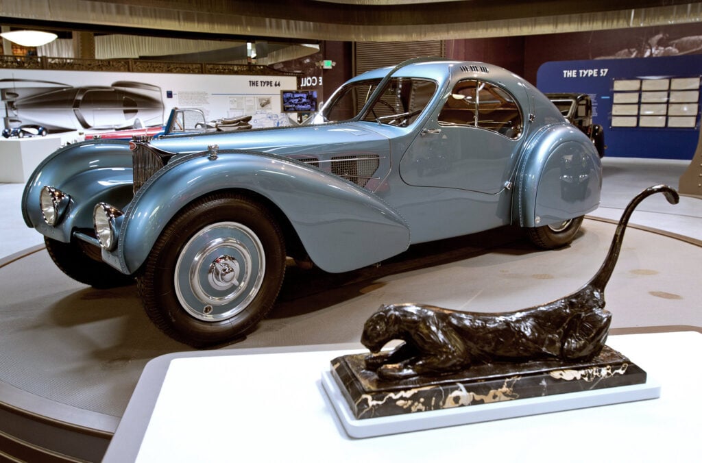 Vintage Bugatti Type 57 blue coupe in museum exhibit, art-deco styling, bronze panther sculpture in foreground.