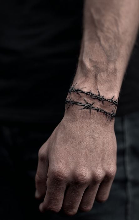 Close-up of male wrist featuring realistic double barbed-wire tattoo wrapped like a bracelet, visible veins, dark background.