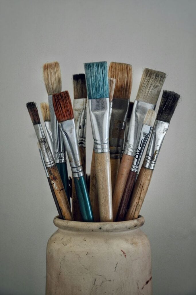 Assorted used artist paintbrushes with stained wooden handles and varied bristles standing in a ceramic jar on a neutral background