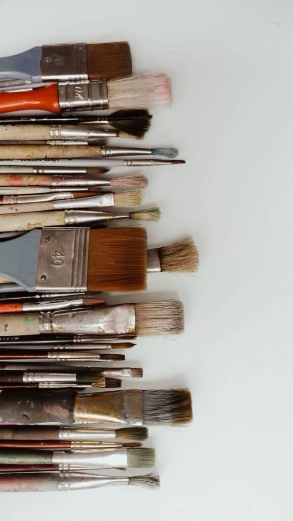 Flatlay of assorted used artist paintbrushes in various sizes and shapes with stained bristles on a white background