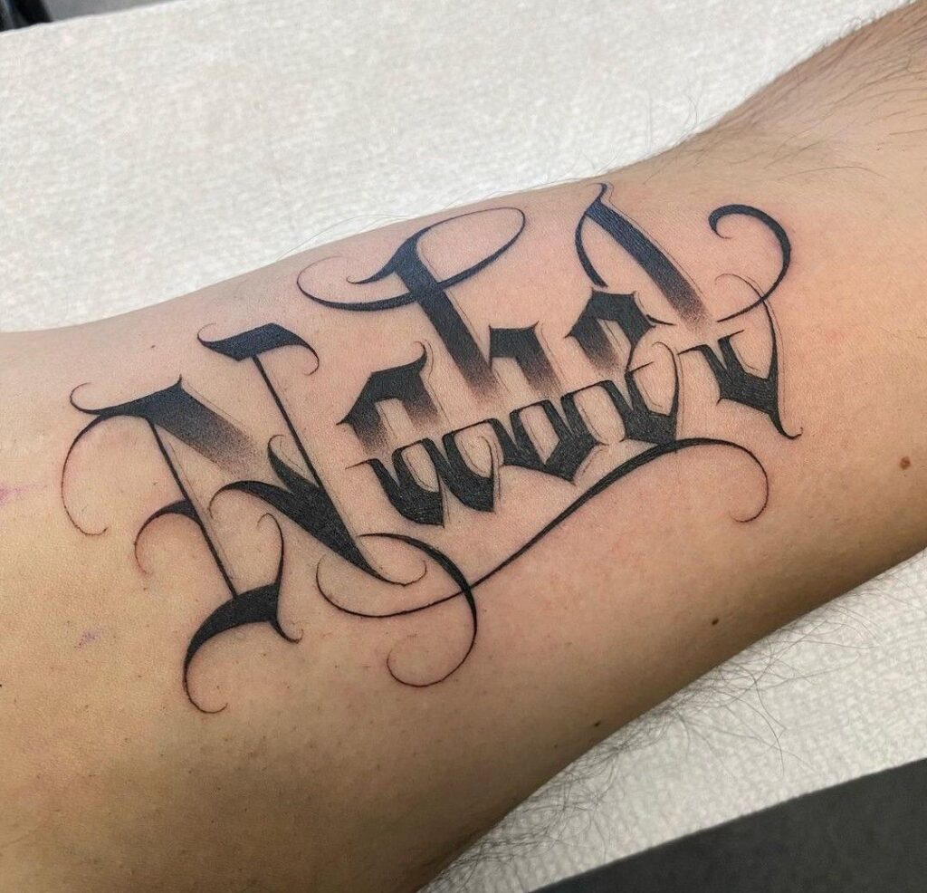 Close-up of black gothic script name tattoo on inner forearm, ornamental flourishes, bold shading and delicate calligraphic swirls.