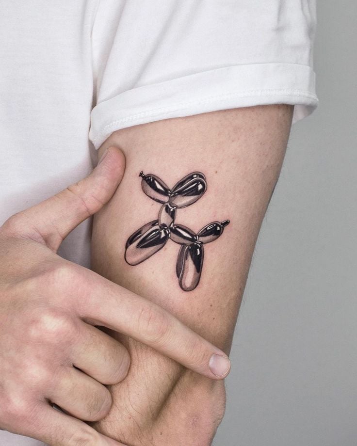 Metallic 3D balloon dog tattoo in shiny black on inner forearm, close-up of arm and hand pointing at design