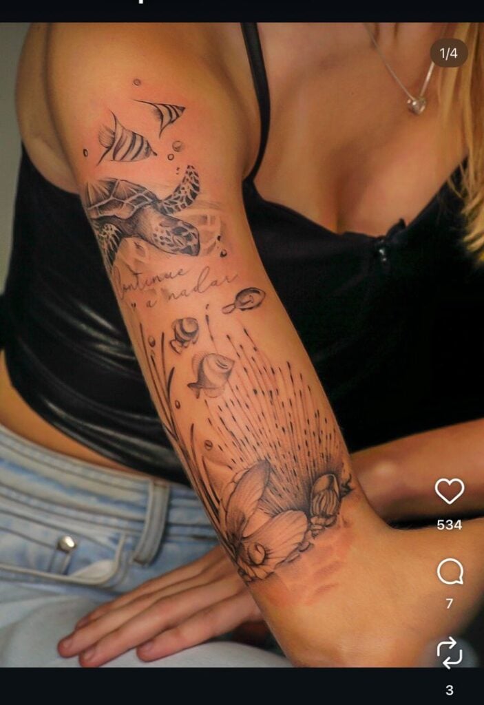 Forearm black-and-grey tattoo of a sea turtle, small fish, bubbles, flowers and cursive script on a woman's arm