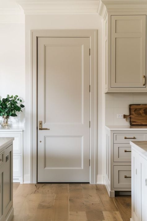 Bright modern kitchen with white paneled door, brass hardware, light shaker cabinets, oak floors and a vase of greenery.