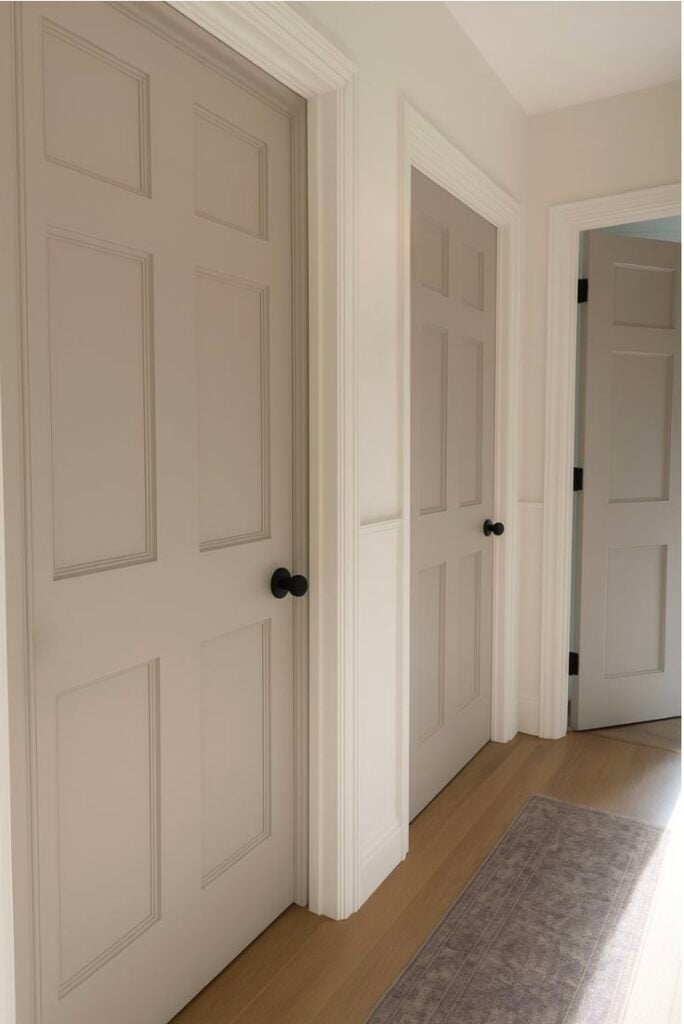 Bright interior hallway with three beige paneled doors, black knobs, white trim, hardwood floor and runner rug.