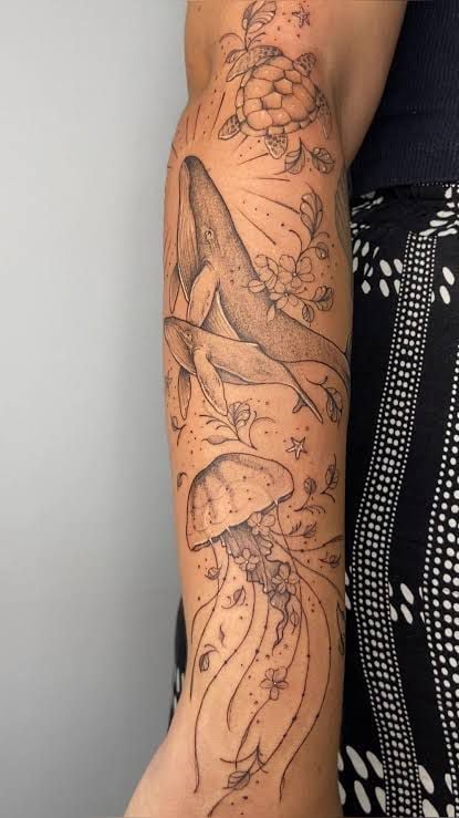 Forearm black-ink marine tattoo featuring humpback whales, jellyfish, a sea turtle and floral dotwork accents extending along the arm.