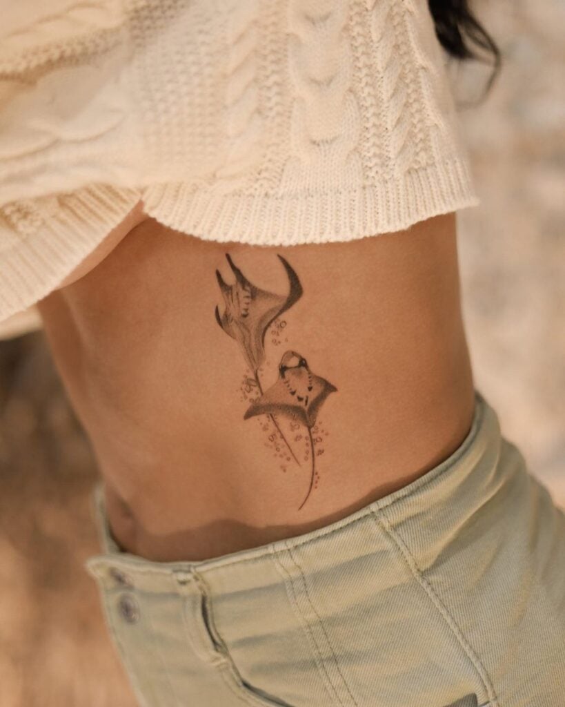Close-up of a person's side torso showing two delicate black-ink manta ray tattoos with bubbles, cream knit top and light denim jeans.