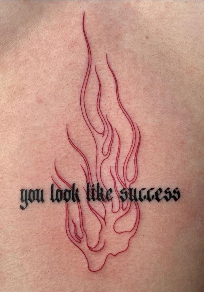 Tattoo close-up: gothic black script reading 'you look like success' over red flame outline on pale skin.
