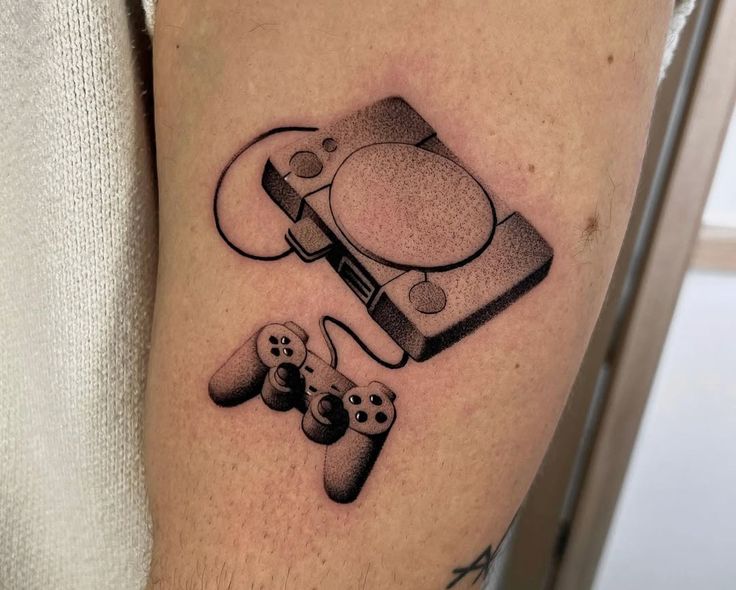 Dotwork blackwork tattoo of a PlayStation PS1 console and DualShock controller on inner thigh, retro gaming ink with stippling shading.