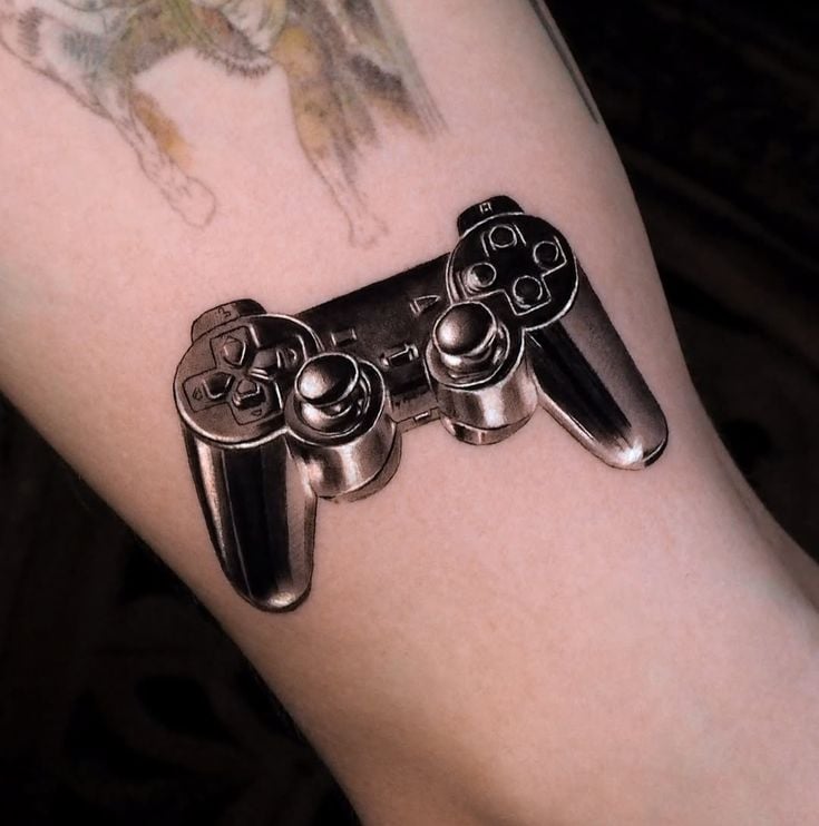 Realistic 3D gaming controller tattoo on forearm, photorealistic black dual-analog design with buttons and shading for depth