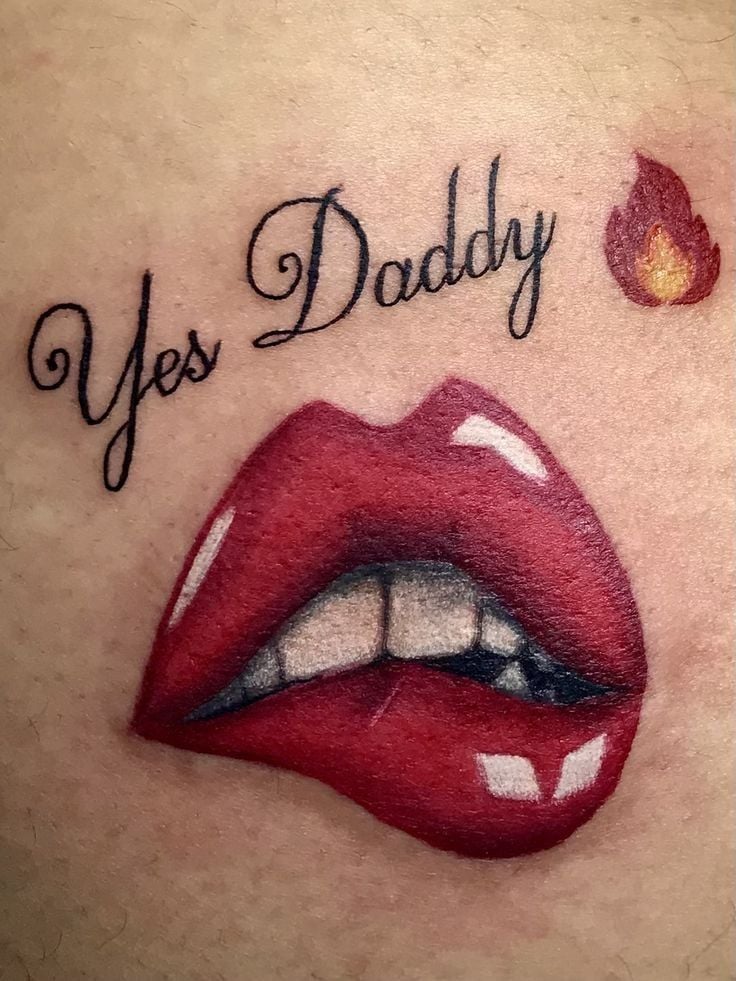 Close-up tattoo: glossy red bitten lips showing teeth, cursive 'Yes Daddy' text and small flame icon on skin.