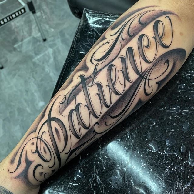 Black-and-gray ornamental script tattoo reading Patience across a forearm, bold lettering with shading and decorative flourishes
