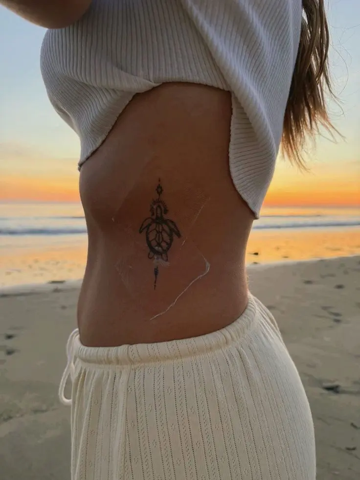 Turtle ribcage tattoo on woman's side at sunset beach, wearing a white crop top and cream knit pants