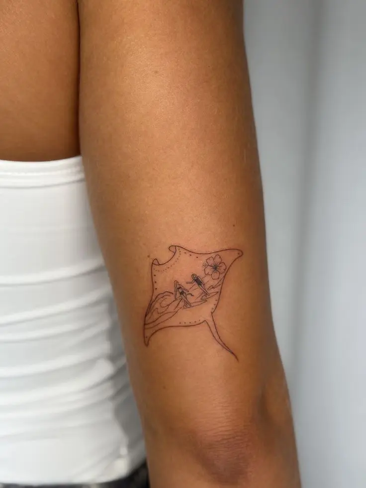 Minimal line-art manta ray tattoo on upper arm featuring tiny surfer riding waves and hibiscus flower