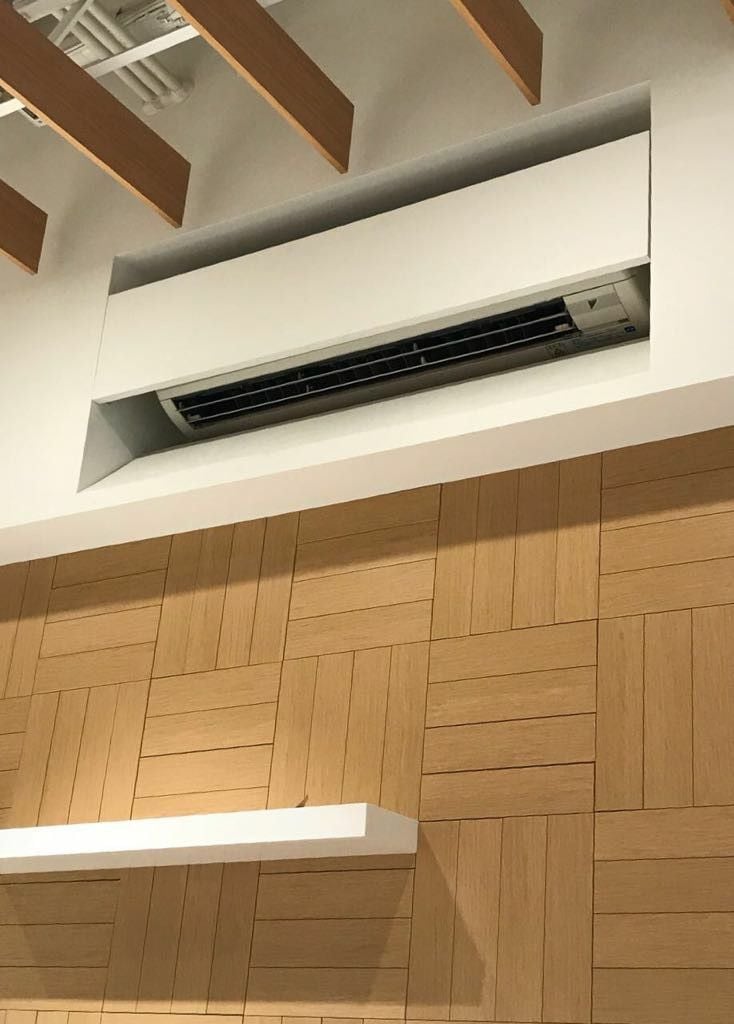 Recessed wall air conditioning unit above wood-paneled accent wall with floating white shelf and exposed ceiling beams in modern interior
