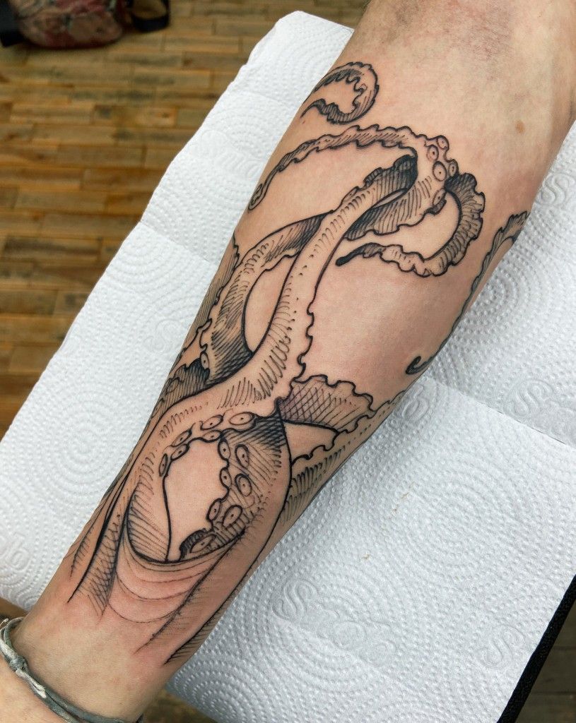 Forearm octopus tattoo in black ink, detailed tentacles and linework, photographed on paper towel with wooden floor background.