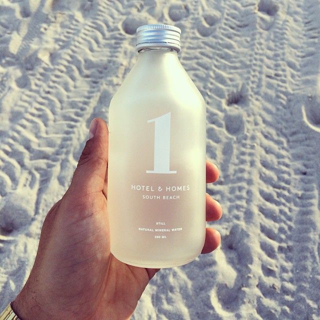 Hand holding frosted glass bottle of 1 Hotel & Homes South Beach mineral water on sunlit sandy beach