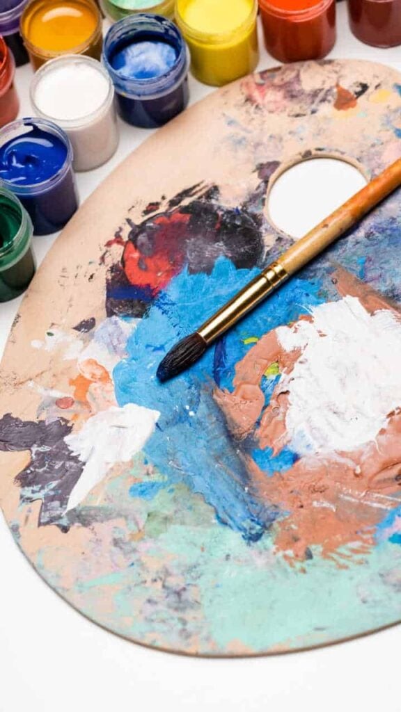 Artist palette with mixed acrylic paints, paintbrush and open paint jars — colorful painting supplies for artists.