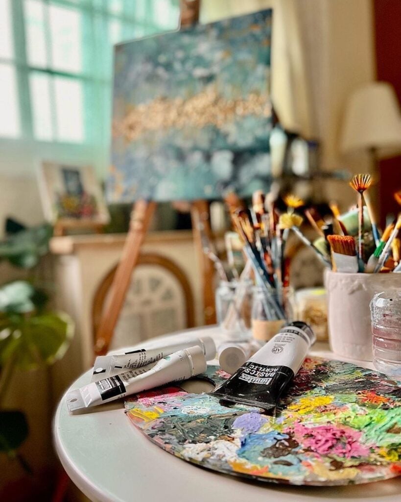 Artist studio close-up: colorful paint palette, acrylic paint tubes and brushes with blurred easel and canvas in background