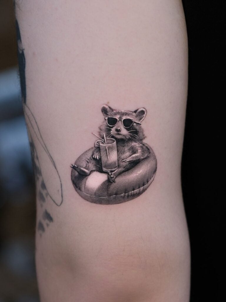 Black and gray inner forearm tattoo of a sunglasses-wearing raccoon lounging on an inflatable tube, holding a cocktail