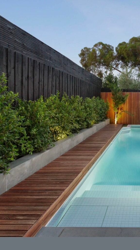 Modern backyard pool with teak deck and tiled steps, raised shrub planter, black wood privacy fence and subtle landscape lighting.