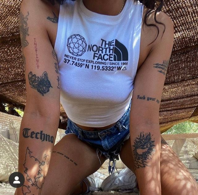 Person in white The North Face crop top and denim shorts, multiple arm and leg tattoos, kneeling in sunlit outdoor woven canopy setting