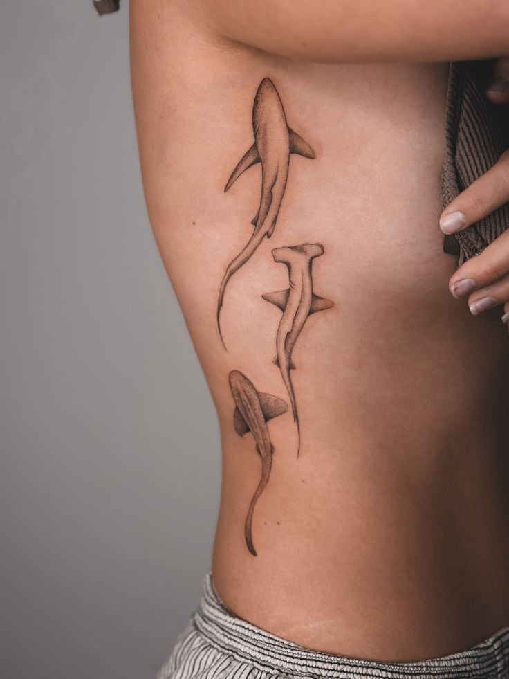 Three minimalist hammerhead shark tattoos flowing down a person's ribcage, fine-line black ink design on bare torso