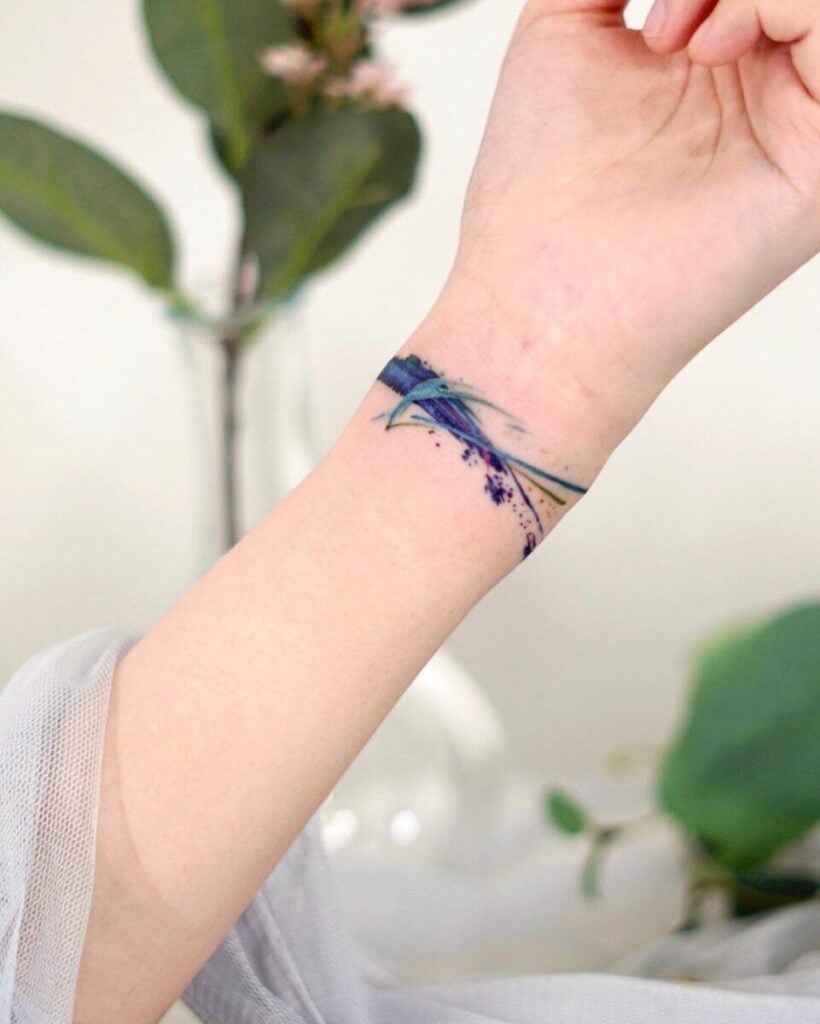 Close-up wrist with blue-purple watercolor feather bracelet tattoo wrapped like a horizontal band on forearm, blurred plant background